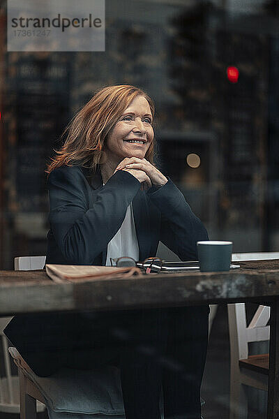 Lächelnde ältere Geschäftsfrau  die mit der Hand am Kinn am Tisch im Café sitzt