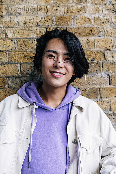 Lächelnder junger Mann mit Kapuzenjacke vor einer Backsteinmauer