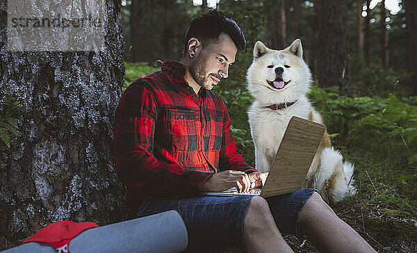 Mittlerer erwachsener Mann  der einen Laptop benutzt  während er mit einem Hund im Wald sitzt