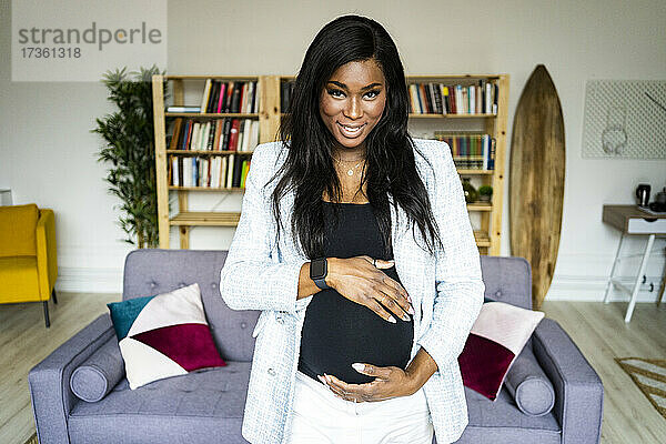Lächelnde junge schwangere Geschäftsfrau  die ihren Unterleib im Wohnzimmer zu Hause berührt