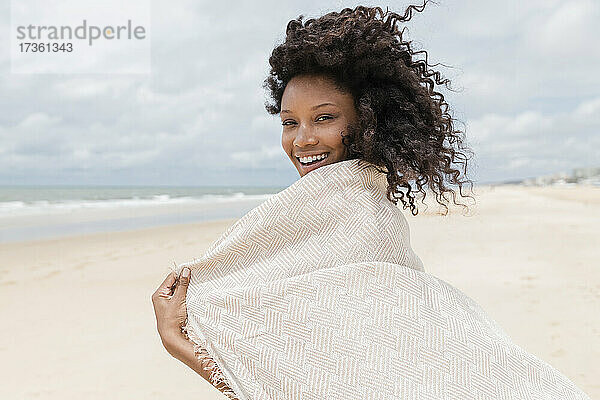 Lächelnde junge Frau mit Handtuch schaut über die Schulter am Strand