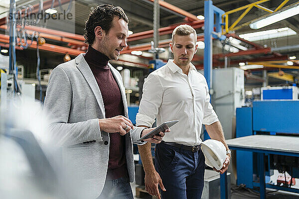 Geschäftsmann  der ein digitales Tablet benutzt  während er an einem männlichen Kollegen in einer Fabrik vorbeigeht