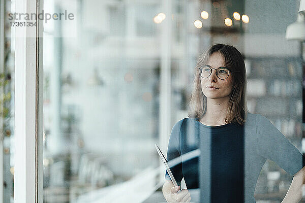 Junge Frau mit Brille  stehend mit Laptop  gesehen durch ein Glasfenster