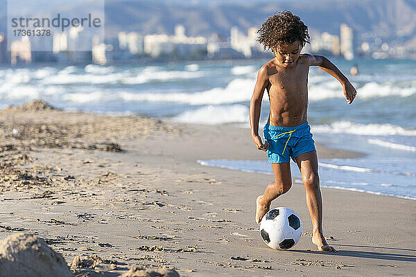 Hemdloser Junge spielt mit Fußball am Strand