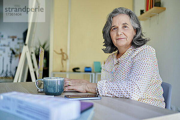 Ältere Geschäftsfrau mit Kaffeetasse und Laptop am Schreibtisch sitzend
