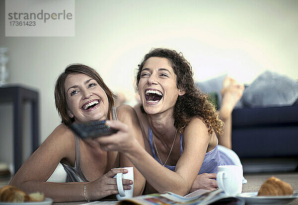 Fröhliche Frau  die mit der Fernbedienung den Sender wechselt  während sie mit einer Freundin zu Hause fernsieht