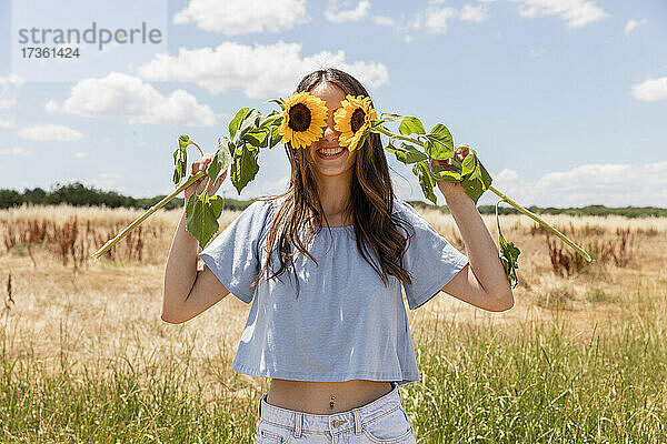 Frau bedeckt Augen mit Sonnenblumen  während sie auf einer Wiese steht