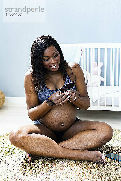Lächelnde schwangere Frau  die im Schneidersitz auf einem Teppich im Schlafzimmer sitzt und sich Ultraschallbilder ansieht