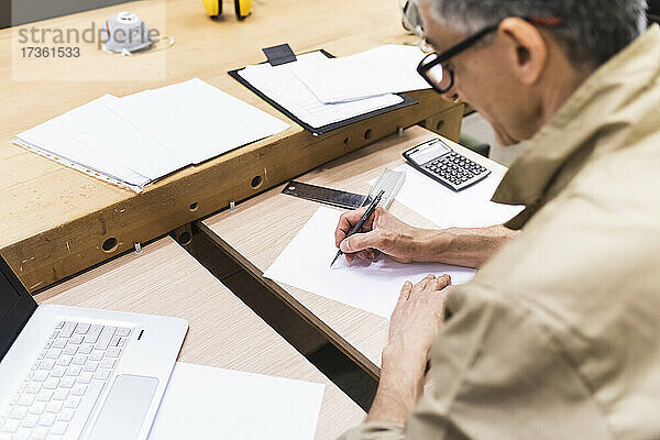 Älterer Mann schreibt am Tisch in einer Werkstatt auf Papier