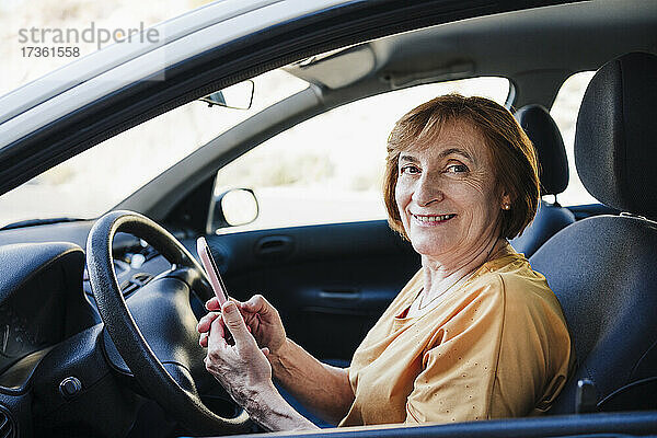 Lächelnde ältere Frau hält ihr Smartphone im Auto sitzend