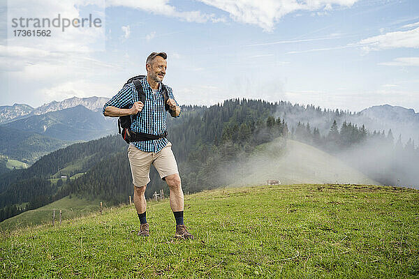 Lächelnder Mann mit Rucksack beim Wandern auf einer Wiese bei nebligem Wetter