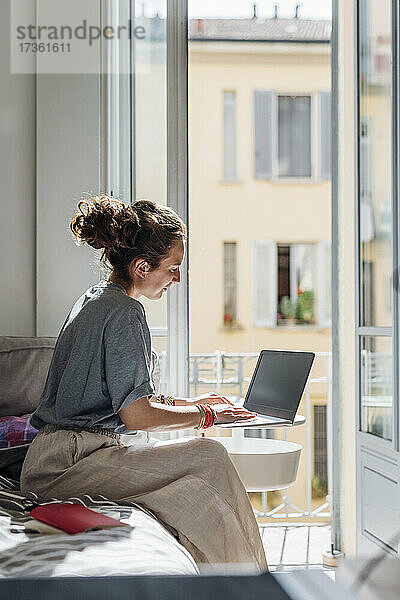 Weibliche Berufstätige  die einen Laptop benutzen  während sie zu Hause auf dem Bett im Sonnenlicht sitzen