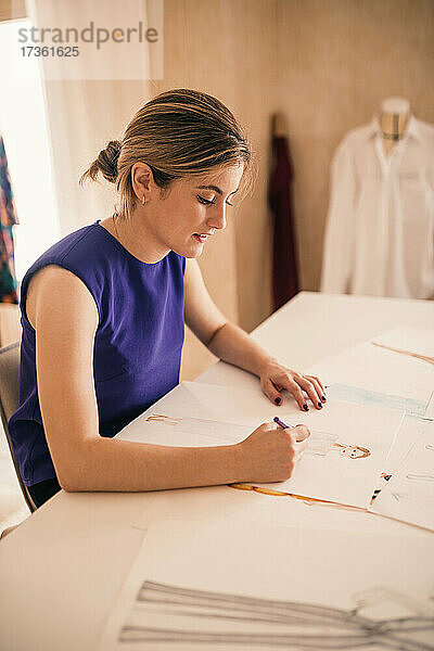 Weibliche Modedesignerin zeichnet auf Papier  während sie an der Werkbank sitzt
