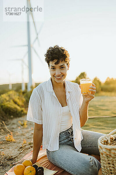 Lächelnde Frau mit Saft in der Hand  die auf einer Picknickdecke in der Nähe von Windrädern sitzt