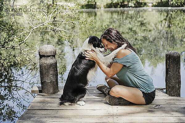 Lächelnde Frau  die ihren Hund umarmt  während sie auf einem Steg am See im Wald sitzt