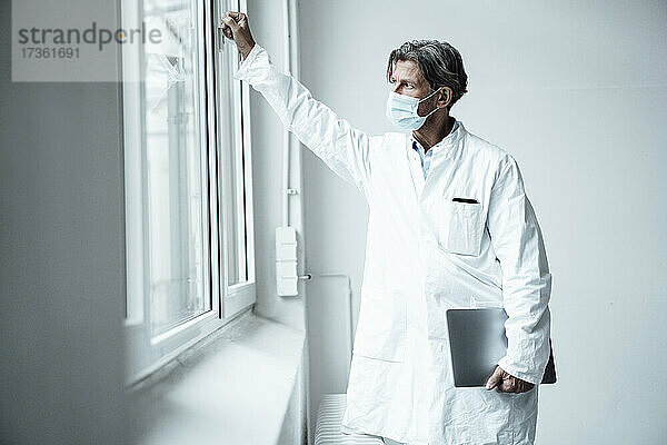 Reifer männlicher Arzt mit Gesichtsschutzmaske schaut durch ein Fenster im Krankenhaus