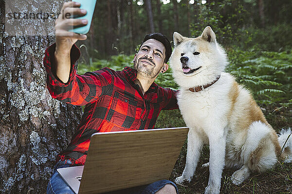 Mann nimmt Selfie mit Hund durch Handy beim Sitzen im Wald