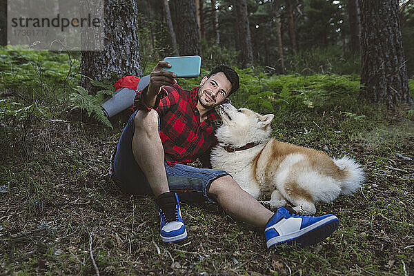 Lächelnder männlicher Wanderer  der ein Selfie mit seinem Haustier über sein Smartphone macht  während er im Wald sitzt
