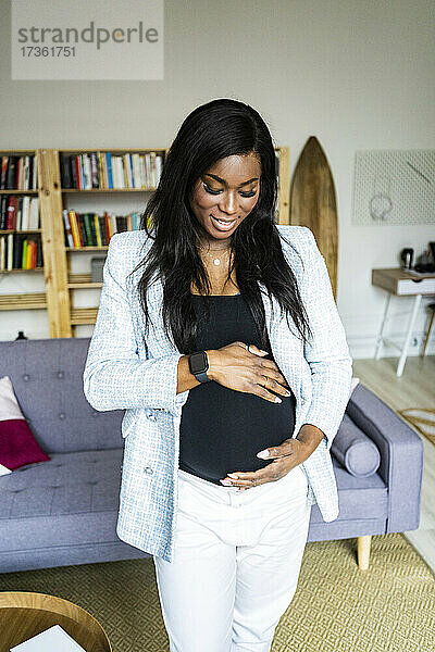 Glückliche junge schwangere Freiberuflerin  die ihren Unterleib berührt  während sie zu Hause steht
