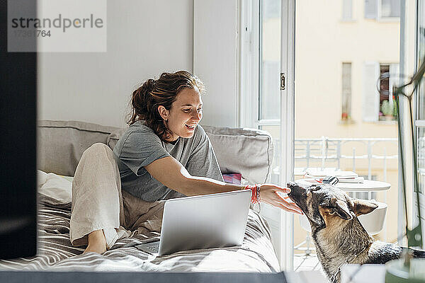 Glückliche Freiberuflerin berührt Hund  während sie auf dem Bett sitzt