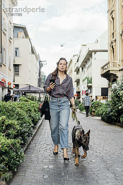 Frau hält Handy in der Hand  während sie mit ihrem Hund auf dem Gehweg spazieren geht