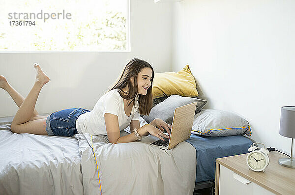 Junge Frau  die einen Laptop benutzt  während sie zu Hause auf dem Bett im Schlafzimmer liegt