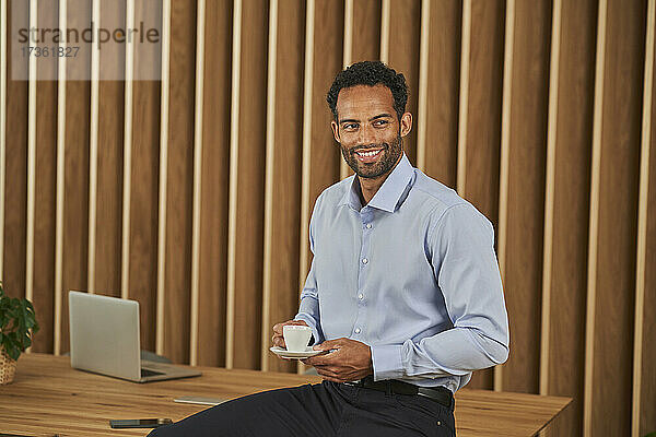 Lächelnder Geschäftsmann hält Kaffeetasse  während er im Büro sitzt