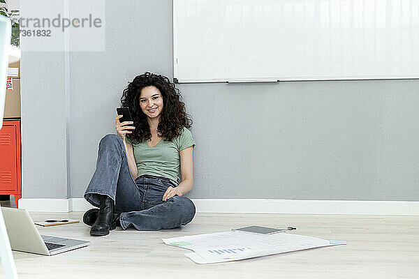 Lächelnde Geschäftsfrau mit Smartphone an die Wand gelehnt  während sie im Büro auf dem Boden sitzt