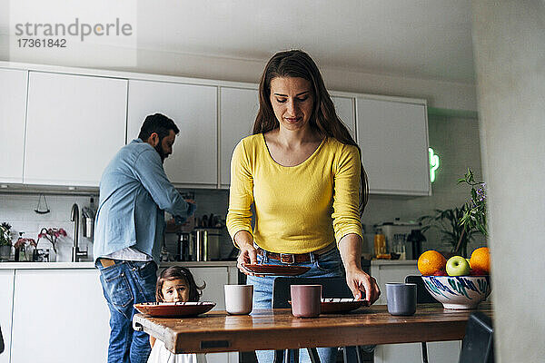 Frau reinigt Tisch  während Familie im Hintergrund zu Hause
