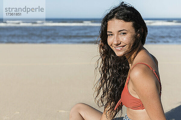 Lächelnde junge Frau am Strand sitzend im Urlaub