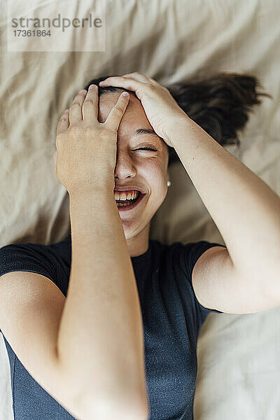 Glückliches Mädchen  das sein Gesicht bedeckt  während es zu Hause auf dem Bett liegt