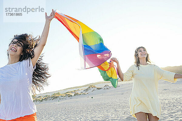 Fröhliche junge Frauen halten die Regenbogenflagge  während sie am Strand aufwachen