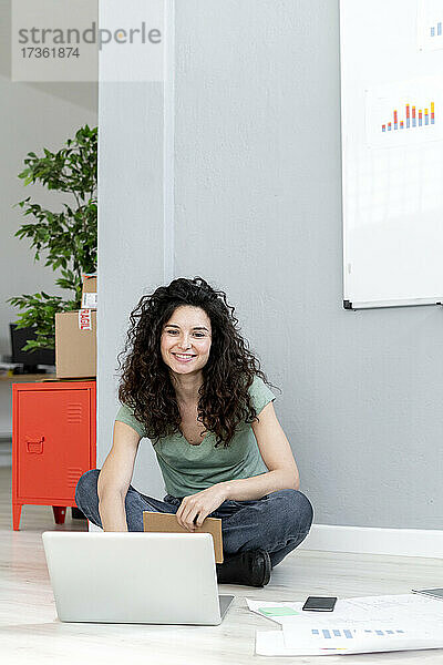 Lächelnde Geschäftsfrau sitzt im Schneidersitz auf dem Boden im Büro