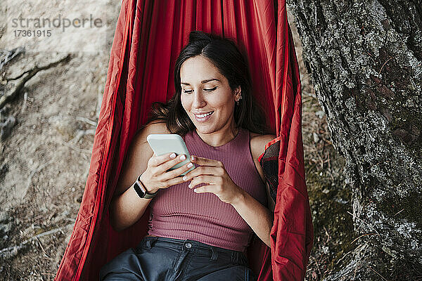 Lächelnde junge Frau  die ein Mobiltelefon benutzt  während sie sich in der Hängematte entspannt