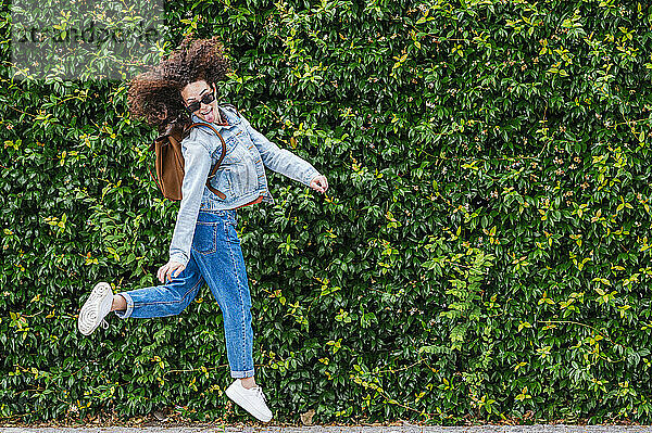 Aufgeregte junge Frau mit Sonnenbrille  die vor grünen Efeupflanzen springt