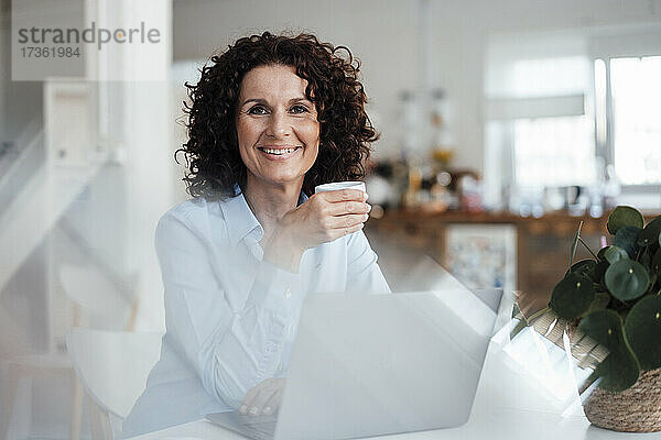 Reife Geschäftsfrau lächelt  während sie mit einer Kaffeetasse am Schreibtisch im Büro sitzt