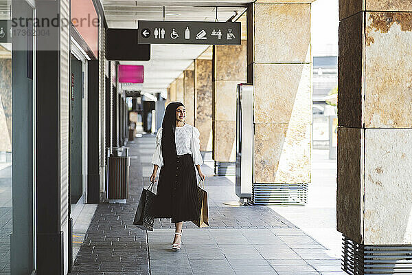Lächelnde Frau mit Einkaufstüten  die beim Gehen auf dem Fußweg wegschaut