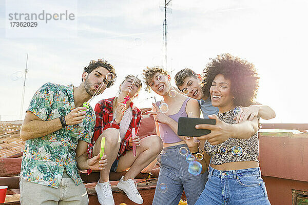 Fröhliche multiethnische Freunde machen ein Selfie beim Spielen mit Seifenblasen auf der Terrasse