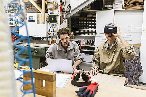 Ein männlicher Schreiner benutzt einen Virtual-Reality-Simulator  während sein Kollege in der Werkstatt einen Laptop benutzt.