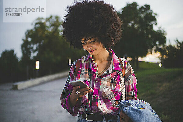 Junge Frau mit Afrofrisur  die in der Abenddämmerung im Freien ihr Smartphone benutzt