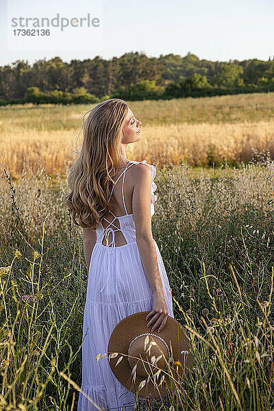 Junge Frau mit Hut in weißem Kleid steht auf einem Feld