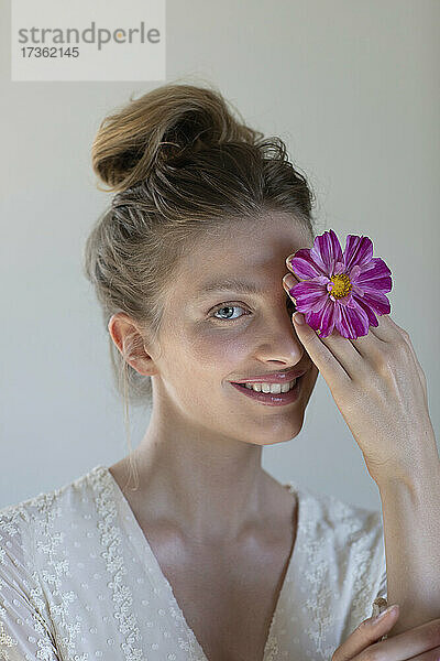 Lächelnde junge Frau bedeckt ihr Auge mit einer lila Blume im Atelier