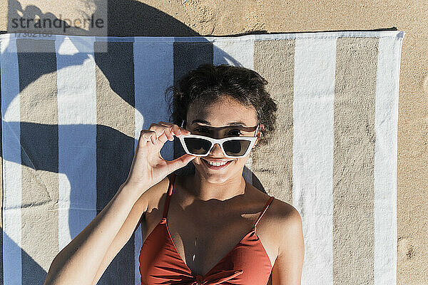 Lächelnde junge Frau mit Sonnenbrille auf einem Handtuch am Strand liegend