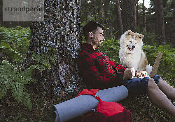 Mann mit Kaffeetasse  der einen Laptop benutzt  während er neben einem Hund im Wald sitzt