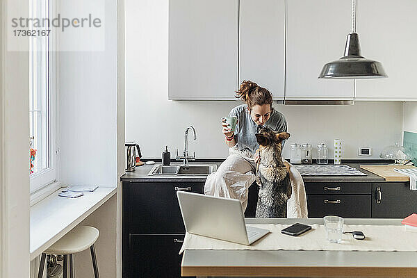 Weibliche Fachkraft spielt mit Hund  während sie in der Kaffeepause auf dem Küchentisch sitzt