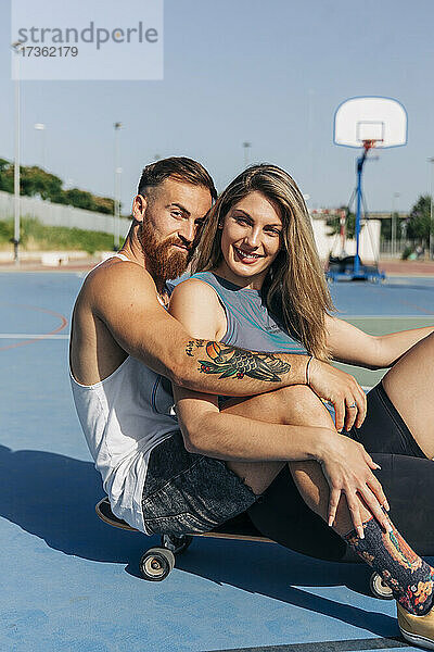 Lächelnde junge Frau sitzt mit ihrem Freund auf dem Skateboard am Basketballplatz