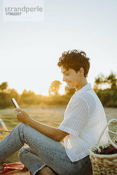 Lächelnde Frau mit Smartphone beim Picknick bei Sonnenuntergang