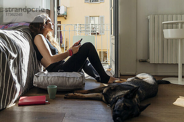 Frau hält Mobiltelefon  während sie mit Hund im Schlafzimmer sitzt