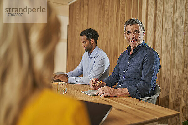 Männlicher Berufstätiger im Gespräch mit einer Mitarbeiterin  während er neben einem Geschäftsmann im Sitzungssaal sitzt