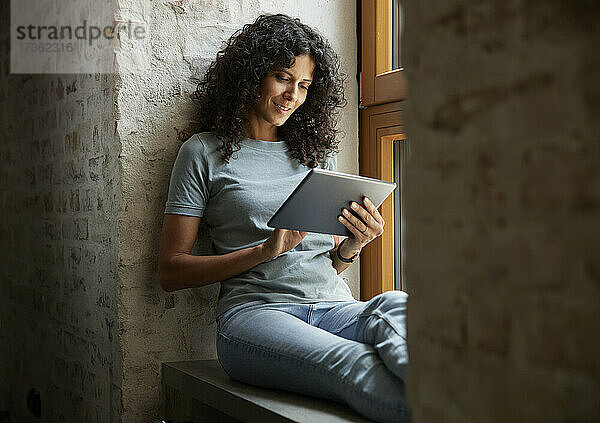 Geschäftsfrau  die ein digitales Tablet benutzt  während sie auf der Fensterbank am Arbeitsplatz sitzt